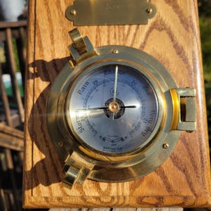 May include: A brass nautical barometer with a round face and a wooden frame. The face has a needle pointing to the "Change" section of the dial, which is marked with "Stormy", "Rain", "Change", "Fair", and "Very Dry".