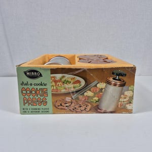 May include: A Mirro Dial-a-Cookie cookie press in its original cardboard box. The packaging features the text Mirro, dial-a-cookie, and cookie press, with illustrations of a metal press and various shaped cookies on a baking sheet.