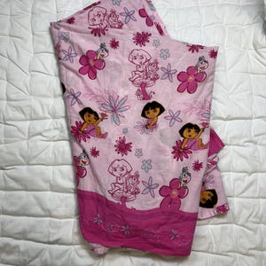May include: Pink Dora the Explorer bed sheet with a repeating pattern of Dora, flowers, and other characters. The sheet has a dark pink border with floral designs. The overall design is bright and colorful.