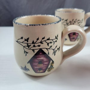 May include: Two ceramic mugs with a beige base color. Each mug features a birdhouse design with a branch and leaves, a small bird, and a blue rim. The handles are a matching beige color.