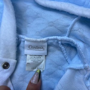 Puede incluir: Prenda de beb&eacute; azul claro con ribete y capucha blancos. La etiqueta dice "Carter's" e incluye informaci&oacute;n de talla: Peque&ntilde;o (0/3m) hasta 5,0 kg. La prenda tiene un cierre de bot&oacute;n a presi&oacute;n. La etiqueta tambi&eacute;n incluye el n&uacute;mero 3523582.