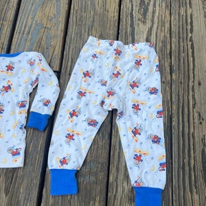 May include: White pajama set with a repeating pattern of astronauts, planets, and moons. The long-sleeved top and pants have blue cuffs. The pajamas are made of a soft, lightweight material.