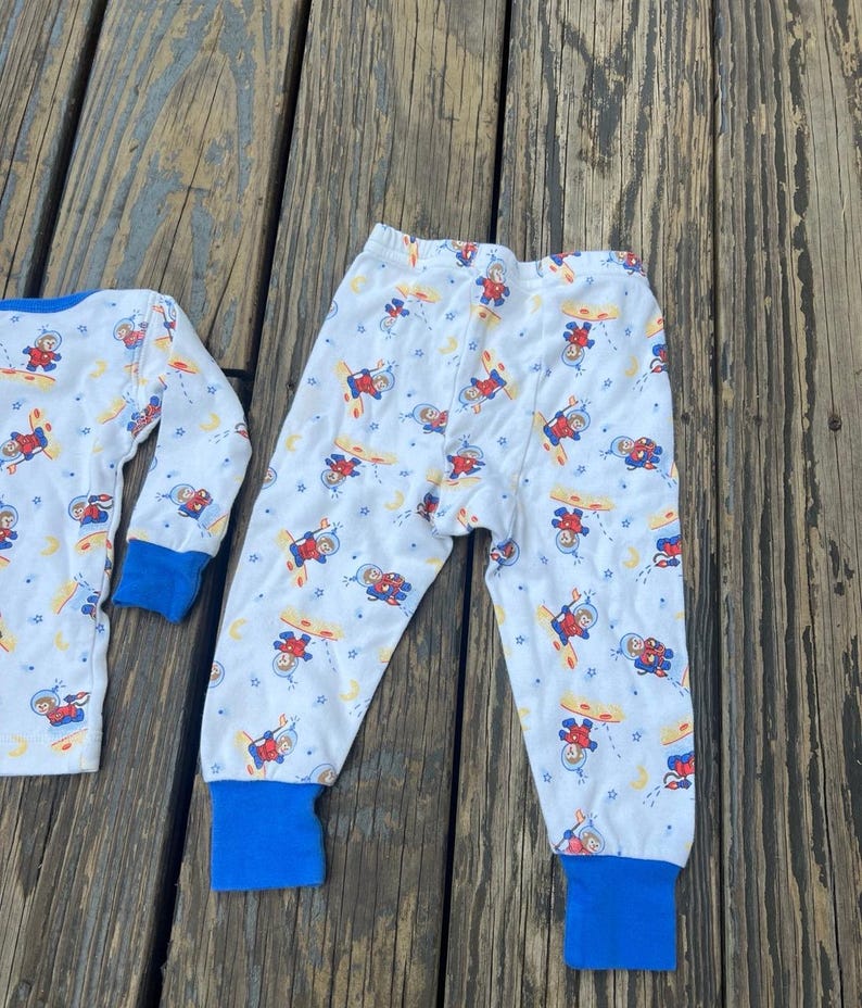 May include: White pajama set with a repeating pattern of astronauts, planets, and moons. The long-sleeved top and pants have blue cuffs. The pajamas are made of a soft, lightweight material.