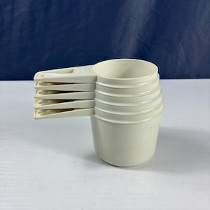 May include: A set of off-white measuring cups. The cups are stacked together, with handles extending to the left. The cups are made of a durable plastic material, ideal for measuring dry and liquid ingredients in the kitchen.