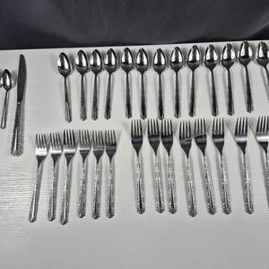 May include: A complete set of stainless steel silverware, including spoons, forks, and knives, arranged on a white surface. The silverware has a decorative pattern on the handles. The set includes various sizes of spoons and forks.