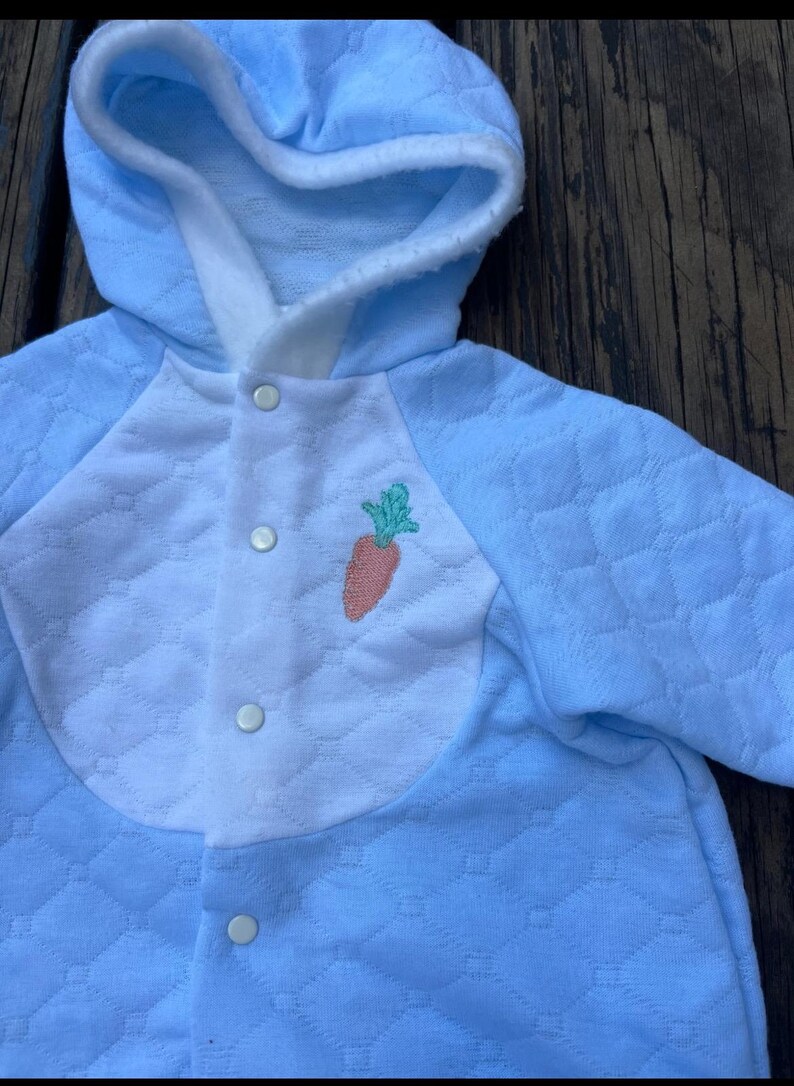 Puede incluir: Mono de beb&eacute; azul claro con capucha y patr&oacute;n acolchado. La parte delantera presenta un panel blanco tipo babero con una aplicaci&oacute;n de zanahoria. El mono tiene tres cierres de bot&oacute;n y est&aacute; dise&ntilde;ado para beb&eacute;s.