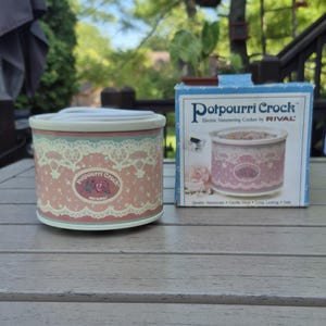 May include: A round Rival Potpourri Crock electric simmering cooker with a pink and cream lace pattern and a rose graphic. The cooker is accompanied by its original blue and white cardboard box featuring the product name and a rose illustration.