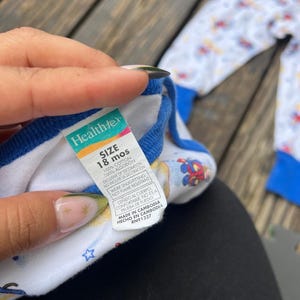 May include: White and blue baby pajamas with a colorful print of cartoon characters. The pajamas have blue trim and a tag that reads "Healthtex" and "SIZE 18 mos". The pajamas are made of 100% cotton.