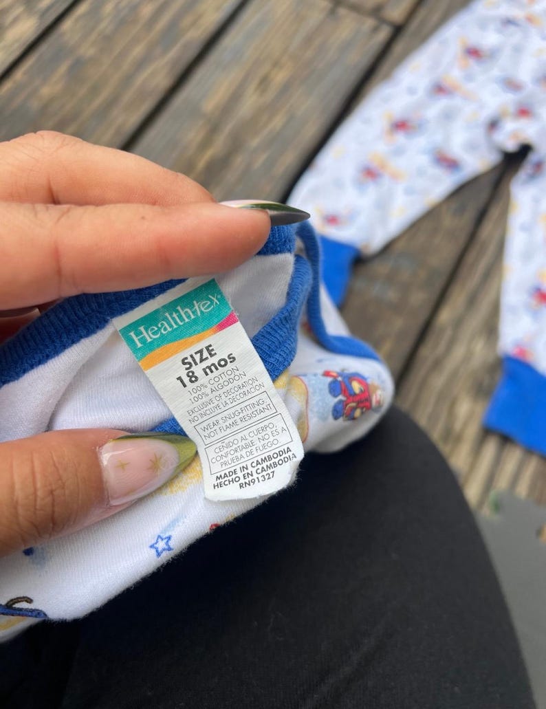 May include: White and blue baby pajamas with a colorful print of cartoon characters. The pajamas have blue trim and a tag that reads "Healthtex" and "SIZE 18 mos". The pajamas are made of 100% cotton.