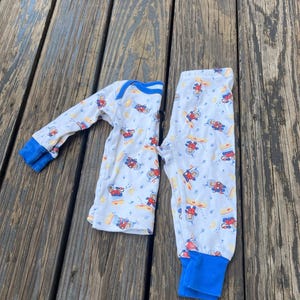 May include: White pajama set with a pattern of astronauts, planets, and stars. The long-sleeved top and pants have blue cuffs and trim. The pajamas are laid out on a weathered wooden surface.