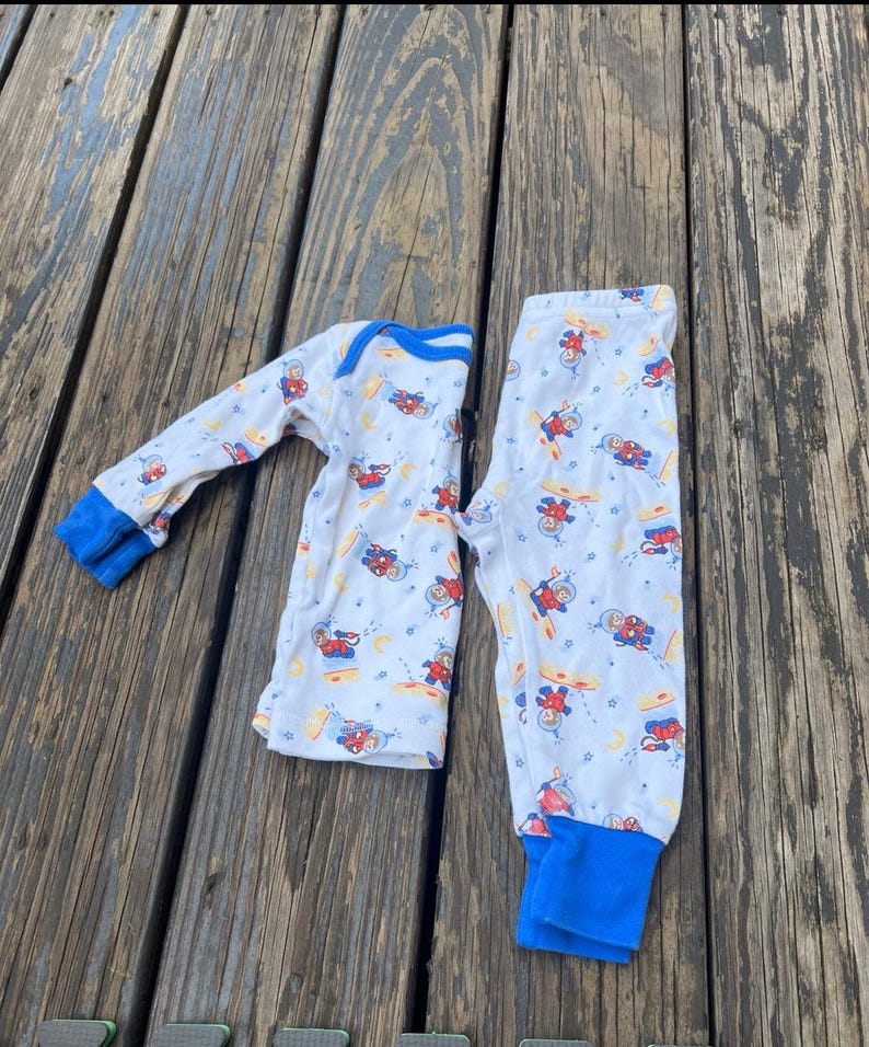 May include: White pajama set with a pattern of astronauts, planets, and stars. The long-sleeved top and pants have blue cuffs and trim. The pajamas are laid out on a weathered wooden surface.