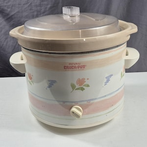 May include: A Rival Crock-Pot slow cooker with a clear lid and cream-colored handles. The white body features a floral pattern with pink, blue, and green accents. The control knob is set to the low position.