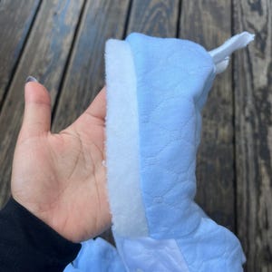 Puede incluir: Un pelele de beb&eacute; azul claro con un ribete blanco y esponjoso. El pelele tiene un patr&oacute;n acolchado y un peque&ntilde;o dise&ntilde;o de fresa bordada. La capucha est&aacute; levantada y el pelele se sostiene en una mano.