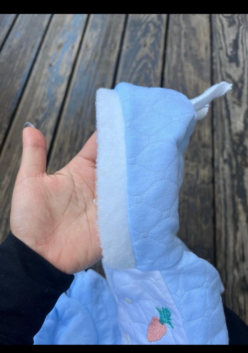 Puede incluir: Un pelele de beb&eacute; azul claro con un ribete blanco y esponjoso. El pelele tiene un patr&oacute;n acolchado y un peque&ntilde;o dise&ntilde;o de fresa bordada. La capucha est&aacute; levantada y el pelele se sostiene en una mano.
