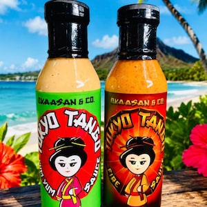 May include: Two 355 ml bottles of Okaasan & Co. Tokyo Tango sauce. The left bottle has a green label with Yum text, and the right bottle has a black and red label with Fire text. Both feature a cartoon character illustration and black caps.