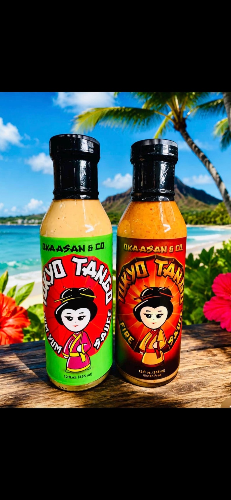 May include: Two 355 ml bottles of Okaasan & Co. Tokyo Tango sauce. The left bottle has a green label with Yum text, and the right bottle has a black and red label with Fire text. Both feature a cartoon character illustration and black caps.