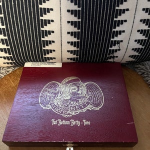 May include: A red wooden box with a gold-colored skull and wings design. The box has a gold-colored latch and the text "Fat Bottom Betty - Toro" is printed on the front.