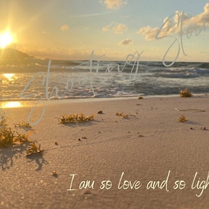 Puede incluir: Una escena de playa con una puesta de sol dorada sobre el océano. La arena es blanca y hay algunas algas en la playa. El texto "I am so love and so light" está escrito en la arena.