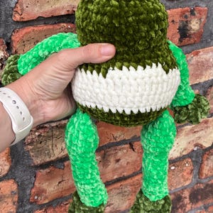 May include: A handmade green and white crochet frog plush toy. The frog has a green head, arms, and legs, with a white band around its midsection. The frog's eyes are black, and the plush is held against a brick wall.