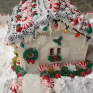 Puede incluir: Una casa de jengibre blanca decorada con bastones de caramelo a rayas rojas y blancas, coronas verdes y luces de colores. El tejado está cubierto de glaseado blanco y caramelos. Una decoración festiva navideña.
