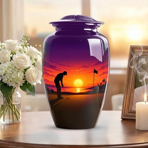 May include: A glossy, dark purple cremation urn featuring a vibrant sunset scene with a silhouette of a golfer on a green, a flagstick, and palm trees. The urn has a rounded lid and a smooth, reflective finish, standing as a decorative vessel.