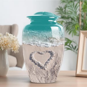 May include: A teal cremation urn with a glossy finish. The urn features a beach scene with a heart made of pebbles on the sand. The top of the urn has a matching teal lid.