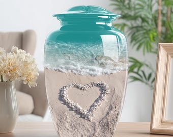 May include: A teal cremation urn with a glossy finish. The urn features a beach scene with a heart made of pebbles on the sand. The top of the urn has a matching teal lid.