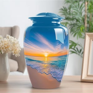 May include: A blue cremation urn with a glossy finish. The urn features a scenic image of a beach at sunset, with the sun setting over the ocean. The urn is sitting on a wooden surface.