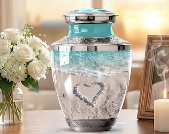 May include: A cremation urn with a turquoise top and silver accents. The clear glass body features a beach scene with a heart in the sand. A bouquet of white roses and a lit candle are in the background.