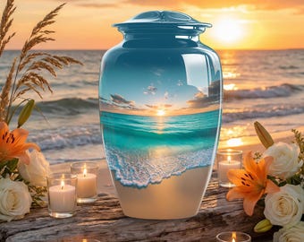 Puede incluir: Una urna funeraria cilíndrica con acabado brillante que muestra una escena impresa de un atardecer sobre un océano turquesa y una playa de arena. La urna tiene una tapa redondeada y está rodeada de rosas blancas, lirios naranjas y velas.