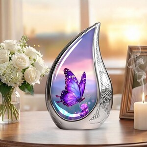 May include: A silver teardrop-shaped urn with a purple butterfly design. The butterfly is set against a sunset backdrop. The urn has intricate floral patterns and is displayed on a wooden surface.
