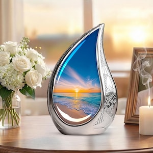 May include: A silver teardrop-shaped urn with a sunset scene inside. The urn has a decorative floral design and sits on a wooden surface. A bouquet of white roses and a lit candle are nearby.