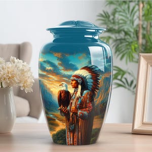 May include: A teal ceramic urn featuring a painting of a Native American figure with an eagle against a sunset landscape. The artwork includes mountains and a colourful sky. The urn has a lid and a glossy finish.