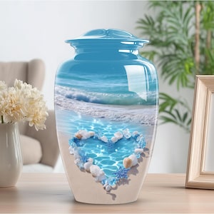 May include: A blue cremation urn with a glossy finish. The urn features a beach scene with a heart-shaped arrangement of shells and blue accents. The urn is sitting on a wooden surface.