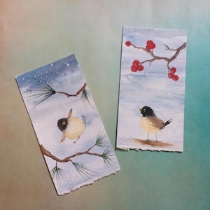 May include: Two watercolor bookmarks featuring birds in a winter scene. One shows a bird perched on a branch with pine needles, the other a bird on a snowy ground with red berries. The backgrounds are light blue with falling snow.