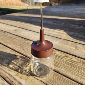 May include: A vintage clear glass and brown plastic kitchen tool. The tool has a brown lid and a white handle. The handle is attached to a metal rod that goes through the lid. The tool is sitting on a wooden deck.