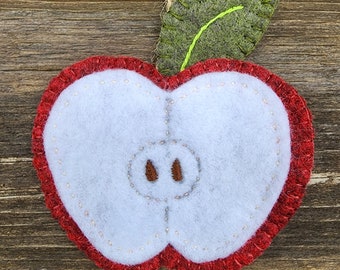 May include: A felt apple ornament featuring a cross-section view with a white center, red outer edge, and a green leaf. The piece is stitched with light-colored thread and includes two brown embroidered seeds, suspended by a thin jute twine loop.