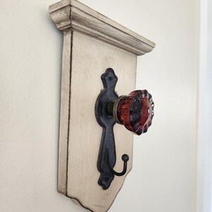 Puede incluir: Un perchero decorativo de pared con un tablero trasero de madera blanquecina envejecida. Cuenta con un gancho de metal negro y un pomo de cristal rojo. El perchero tiene una moldura superior decorativa.