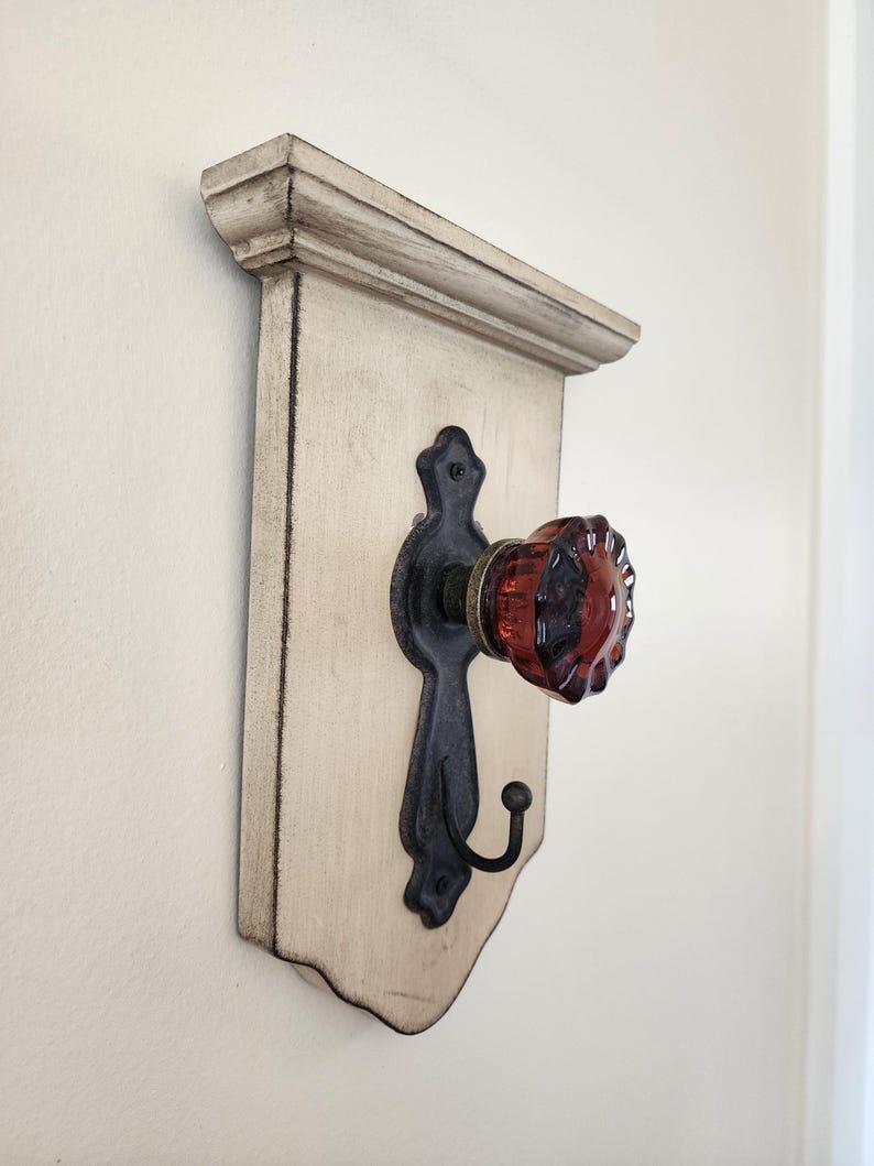 Puede incluir: Un perchero decorativo de pared con un tablero trasero de madera blanquecina envejecida. Cuenta con un gancho de metal negro y un pomo de cristal rojo. El perchero tiene una moldura superior decorativa.