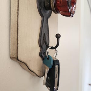 Puede incluir: Un llavero con una base de madera blanquecina envejecida y herrajes de metal oscuro ornamentados. Cuenta con un pomo de puerta de cristal rojo y un gancho que sostiene llaves. El dise&ntilde;o tiene una est&eacute;tica vintage y r&uacute;stica.
