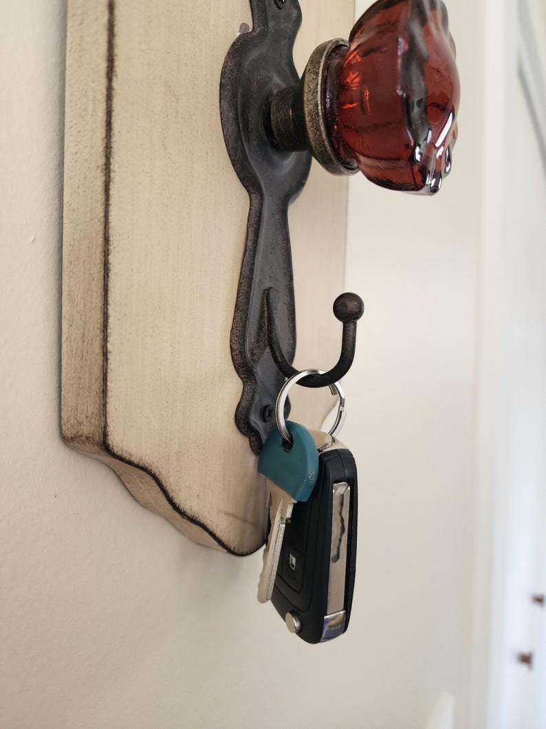 Puede incluir: Un llavero con una base de madera blanquecina envejecida y herrajes de metal oscuro ornamentados. Cuenta con un pomo de puerta de cristal rojo y un gancho que sostiene llaves. El dise&ntilde;o tiene una est&eacute;tica vintage y r&uacute;stica.