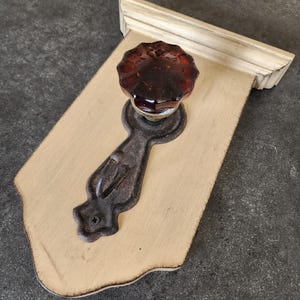 May include: A decorative wall-mounted cabinet knob with a dark amber glass knob and a cast iron backplate. The knob is mounted on a cream-colored wooden plaque with a decorative edge and a white trim.