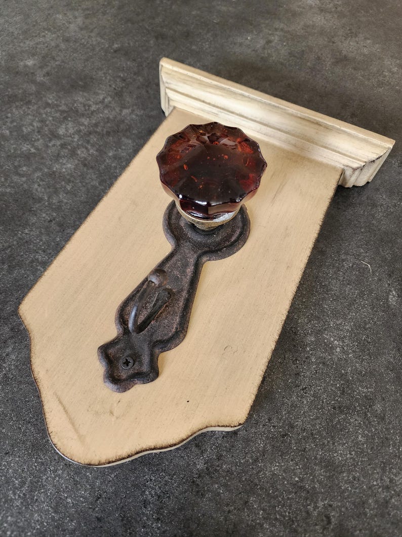 Puede incluir: Pomo decorativo de armario montado en la pared con un pomo de cristal &aacute;mbar oscuro y una placa trasera de hierro fundido. El pomo est&aacute; montado sobre una placa de madera color crema con borde decorativo y ribete blanco.