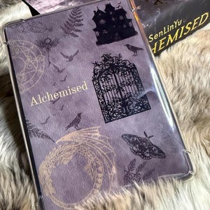 Könnte beinhalten: Eine transparente Schutzhülle mit lila Hintergrund, verziert mit gotischen Illustrationen eines Spukhauses, Tors und einer Krähe. Das Wort "Alchemised" ist in Gold gedruckt. Im Hintergrund ist ein Buch mit dem Titel "Hemised" zu sehen.