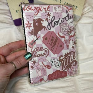 May include: A pink and white phone case with a cowgirl theme. The case features a checkered pattern, a cowboy hat, boots, a skull, and the words "Cowgirl", "Howdy", "Love Coupon", "Yee Haw", "Rodeo", and "Saloon".
