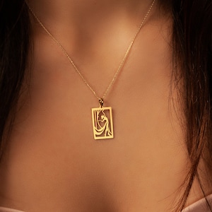 Puede incluir: Un collar de oro con un colgante rectangular que presenta un diseño recortado de una mujer con un vestido fluido.