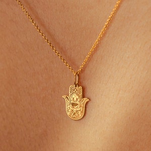 May include: A gold chain necklace with a hamsa hand pendant. The pendant is detailed with an eye in the center and intricate designs.