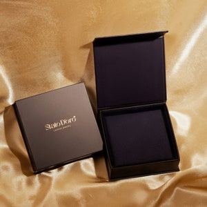May include: Two black boxes with a gold logo that reads "Stato D'oro custom jewelry". The box on the right is open, revealing a black velvet interior.