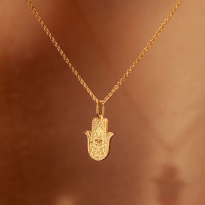 May include: A gold chain necklace with a hamsa hand pendant. The pendant is detailed with an eye in the center and intricate patterns.