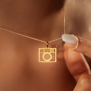 Pode incluir: Um colar de corrente de ouro com um pingente em forma de câmera. O pingente é um contorno simples de uma câmera.